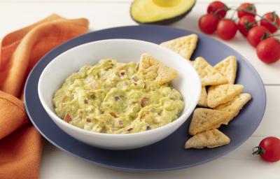 Fotografia em tons de azul em uma bancada de madeira clara com um pano laranja ao lado e um prato grande azul e um prato fundo branco com o guacamole dentro. Ao lado, um pedaço de avocado, alguns tomatinhos e biscoitinhos. Fotografia em tons de azul em uma bancada de madeira clara com um pano laranja ao lado e um prato grande azul e um prato fundo branco com o guacamole dentro. Ao lado, um pedaço de avocado, alguns tomatinhos e biscoitinhos.