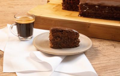 Fotografia de uma bancada de madeira, e sobre ela tem uma torta de chocolate com café em um prato de vidro em tom pastel, que está apoiado sobre um pano branco. Ao lado da sobremesa tem uma xícara com Nescafé, também sobre o pano. Fotografia de uma bancada de madeira, e sobre ela tem uma torta de chocolate com café em um prato de vidro em tom pastel, que está apoiado sobre um pano branco. Ao lado da sobremesa tem uma xícara com Nescafé, também sobre o pano.