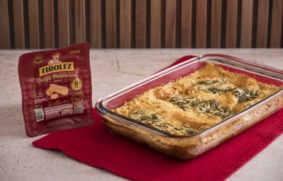 Foto da receita de Lasanha e Queijo Parmesão e Pesto, servida em uma travessa de vidro, sobre um pano vermelho. Ao lado dela há uma embalagem de Queijo Parmesão Tirolez