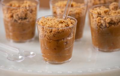 Fotografia em tons de branco com vários potinhos de plástico ao centro. Dentro de cada potinho existe um brigadeiro de cor caramelo feito com paçoca e coberto com paçoca triturada.