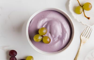 Fotografia em tons de branco e roxo com um pote branco ao centro. Dentro do pote existe uma receita de mousse de uva de cor roxa com algumas uvas verdes de enfeite. Fotografia em tons de branco e roxo com um pote branco ao centro. Dentro do pote existe uma receita de mousse de uva de cor roxa com algumas uvas verdes de enfeite.