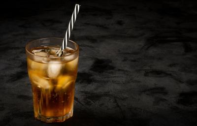 Fotografia em tons escuros de um copo de vidro com uma bebida de guaraná com cubos de gelo e um canudo preto e branco dentro do copo, o qual está sobre uma mesa preta.