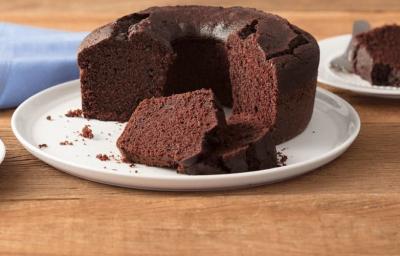 Fotografia em tons de marrom e branco de uma bancada de madeira clara com um paninho azul claro e um prato branco redondo com o bolo de chocolate. Aos lados dois pratos brancos pequenos com fatias de bolo de chocolate e um garfo.