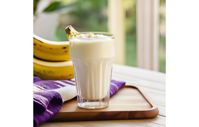 Fotografia em tons de roxo e amarelo com um copo de vidro ao centro. Dentro do copo existe um shake de coco, batata doce e banana de cor branca. Ao fundo existe duas bananas