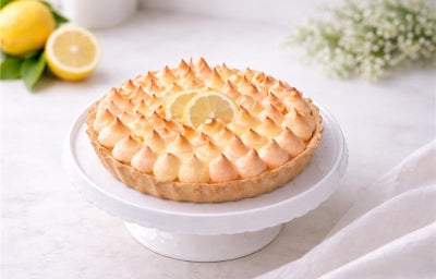 Imagem mostra uma torta de limão em dons alaranjados, sobre um prato de bolo branco em cima de uma mesa com toalha branca.