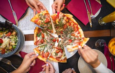Fotografia vista de cima de uma pizza com escarola, linguiça, muçarela de búfala e manjericão com tomates. A pizza está sobre um prato raso branco, cortada em 8 fatias. Na imagem, há quatro pessoas estão puxando uma fatia, e ao redor, copos e talheres. Fotografia vista de cima de uma pizza com escarola, linguiça, muçarela de búfala e manjericão com tomates. A pizza está sobre um prato raso branco, cortada em 8 fatias. Na imagem, há quatro pessoas estão puxando uma fatia, e ao redor, copos e talheres.