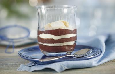 Fotografia em tons de azul e cinza, em uma bancada contém um pano azul e em cima um prato com um copo que contém açaí, banana e leite em pó. Ao lado uma colher para servir.