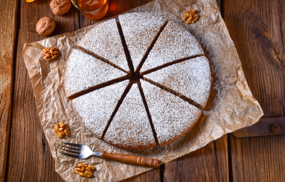 Foto em tons de marrom da receita de bolo alemão de mel servida cortada em 8 pedaços polvilhada com açúcar de confeiteiro, bolo em cima de uma mesa de madeira com nozes ao lado Foto em tons de marrom da receita de bolo alemão de mel servida cortada em 8 pedaços polvilhada com açúcar de confeiteiro, bolo em cima de uma mesa de madeira com nozes ao lado