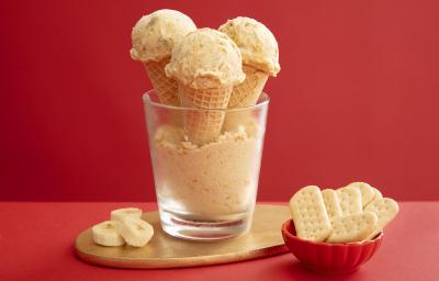 Foto da receita de Sorvete de Banoffee. Observa-se um copo de vidro com farofinha de biscoito no fundo e três casquinhas de sorvete com uma bola dispostas no copo. Ao lado direito, um potinho com biscoitos e, ao esquerdo, rodelas de banana