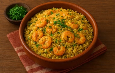 Fotografia de um recipiente marrom de vidro fundo, médio, sobre um pano dobrado de tom rosa e branco apoiado em uma mesa de madeira. Dentro do recipiente tem arroz paraense com camarões por cima e um pouco de cheiro verde salpicado. Fotografia de um recipiente marrom de vidro fundo, médio, sobre um pano dobrado de tom rosa e branco apoiado em uma mesa de madeira. Dentro do recipiente tem arroz paraense com camarões por cima e um pouco de cheiro verde salpicado.
