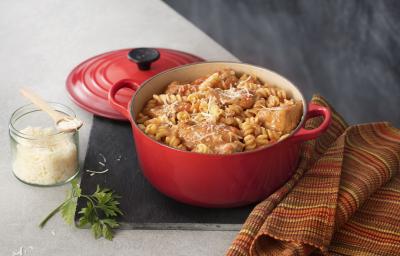 Fotografia de uma panela vermelha aberta com macarrão parafuso, frango e queijo parmesão ralado em cima e ao lado está a tampa da panela vermelha com ponta preta, um guardanapo listrado em tons terrosos e uma pode de vidro transparente com queijo ralado.