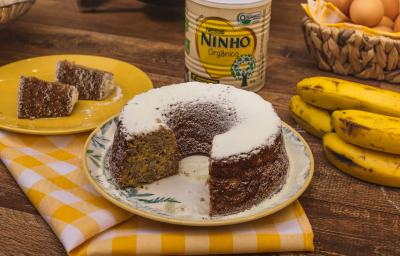 Fotografia em tons de amarelo e marrom de uma bancada de madeira com um pano xadrez amarelo e branco de um bolo de banana polvilhado com NINHO polvilhado. Ao fundo uma lata de NINHO, bananas e ovos. Fotografia em tons de amarelo e marrom de uma bancada de madeira com um pano xadrez amarelo e branco de um bolo de banana polvilhado com NINHO polvilhado. Ao fundo uma lata de NINHO, bananas e ovos.
