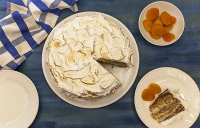 Fotografia em tons de azul e laranja em uma bancada azul, com um pano xadrez azul, um prato branco redondo com a torta de nozes em cima dele. Ao lado, um prato menor com a fatia da torta e damascos ao lado dele. Fotografia em tons de azul e laranja em uma bancada azul, com um pano xadrez azul, um prato branco redondo com a torta de nozes em cima dele. Ao lado, um prato menor com a fatia da torta e damascos ao lado dele.