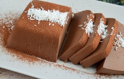 Em cima de uma mesa, prato branco com Bavaroise cortados em fatias, decorado com coco ralado.