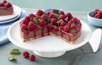 Fotografia em tons de vermelho em uma bancada de madeira azul, um pano azul claro, um suporte para bolo branco com a cheesecake de chocolate com calda de framboesa em cima do suporte, decorado com raspas de chocolate e framboesas. Fotografia em tons de vermelho em uma bancada de madeira azul, um pano azul claro, um suporte para bolo branco com a cheesecake de chocolate com calda de framboesa em cima do suporte, decorado com raspas de chocolate e framboesas.