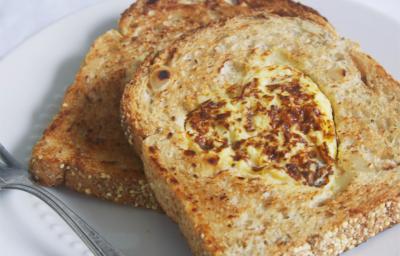 Fotografia em tons de bege em uma mesa de madeira clara com um prato branco raso e duas fatias de pão integral com a omelete dentro dele.