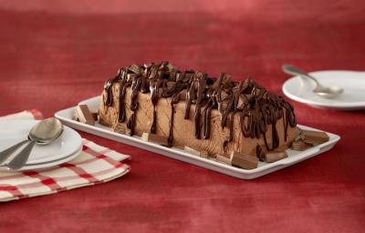 Fotografia em tons de chocolate e vinho, ao centro régua branca contendo torta de chocolate coberta e decorada com chocolate, ao lados pratos com colheres, dois deles sobre guardanapo branco com listras vermelhas, tudo sobre bancada na cor vinho.