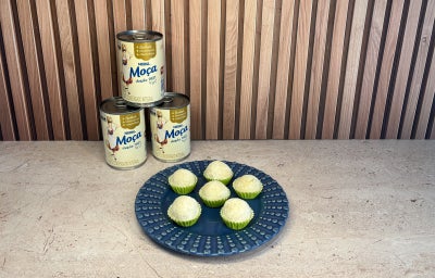 Seis brigadeiros brancos em forminhas verdes sobre prato azul, com três latas de LEITE MOÇA® ao fundo.