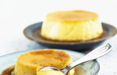 Fotografia de um flan de iogurte e maracujá com calda de maracujá com uma colher de sobremesa cortando um pedaço sobre um prato de tom verde. Ao fundo, outro flan, sobre uma mesa branca.
