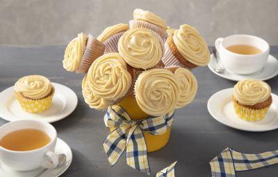 Fotografia em tons de amarelo em uma bancada cinza com um vaso de buquê de cupcake ao centro com um laço xadrez em amarelo e azul. Ao lado, xícaras brancas com chá e pratos brancos com um cupcake em cada um. Fotografia em tons de amarelo em uma bancada cinza com um vaso de buquê de cupcake ao centro com um laço xadrez em amarelo e azul. Ao lado, xícaras brancas com chá e pratos brancos com um cupcake em cada um.