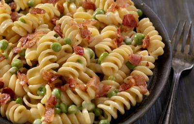 fotografia em tons de preto e bege de uma bancada preta vista de cima. Contém um recipiente preto redondo com macarrão parafuso, pedaços de bacon e ervilhas.