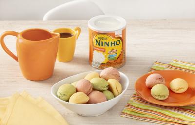 Fotografia em tons de branco e laranja de uma bancada branca com um recipiente branco oval, sobre ele pães de queijo. Ao lado um paninho listrado com um prato laranja com pães de queijo, uma lata de NINHO zero lactose, uma xícara e uma jarra laranjas.