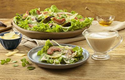Fotografia em tons de verde em uma bancada de madeira com um prato de madeira grande com salada de alface, figo e queijos. Mais à frente, um prato menor com uma porção pequena da salada. Ao lado, um potinho com molho, mel e queijo parmesão.