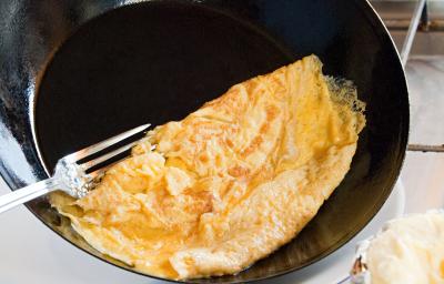 Fotografia de uma omelete dobrada em uma frigideira funda preta que está inclinada para colocar o ovo no prato, e um garfo segundo a omelete. Ao fundo, ovos sobre uma mesa branca. Ao lado do prato que vai receber a omelete, um pano de mesa vermelho.