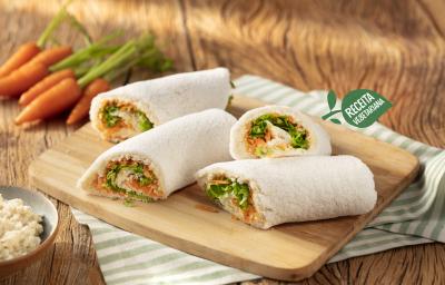 Fotografia em tons de marrom, verde e branco de uma bancada de madeira com um paninho listrado branco e verde, com uma tábua de madeira, sobre ela quatro rolinhos de tapioca. Ao fundo cenouras e ao lado um recipiente pequeno redondo com ricota. Fotografia em tons de marrom, verde e branco de uma bancada de madeira com um paninho listrado branco e verde, com uma tábua de madeira, sobre ela quatro rolinhos de tapioca. Ao fundo cenouras e ao lado um recipiente pequeno redondo com ricota.