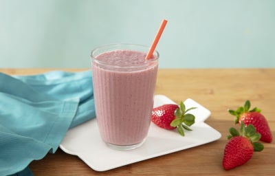 Foto da receita de shake de açaí com novasource proline servida em um copo de vidro alto sobre uma louça branca em cima de uma mesa de madeira com morangos Foto da receita de shake de açaí com novasource proline servida em um copo de vidro alto sobre uma louça branca em cima de uma mesa de madeira com morangos
