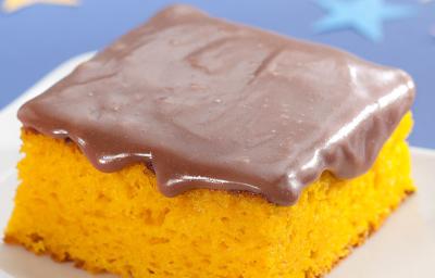 Fotografia em tons de azul, laranja e marrom de uma bancada azul, sobre ela um prato branco quadrado com uma fatia de bolo de cenoura com cobertura de chocolate. Ao lado um garfo verde. Ao fundo uma estrela azul e uma rosa. Fotografia em tons de azul, laranja e marrom de uma bancada azul, sobre ela um prato branco quadrado com uma fatia de bolo de cenoura com cobertura de chocolate. Ao lado um garfo verde. Ao fundo uma estrela azul e uma rosa.