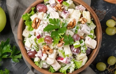 Fotografia de uma salada vibrante em uma tigela de madeira. A mistura inclui cubos de batatas cozidas, maçã-verde, uvas roxas e nozes, coberta com um molho cremoso e decorada com salsa, destacando frescor e texturas harmoniosas. Fotografia de uma salada vibrante em uma tigela de madeira. A mistura inclui cubos de batatas cozidas, maçã-verde, uvas roxas e nozes, coberta com um molho cremoso e decorada com salsa, destacando frescor e texturas harmoniosas.