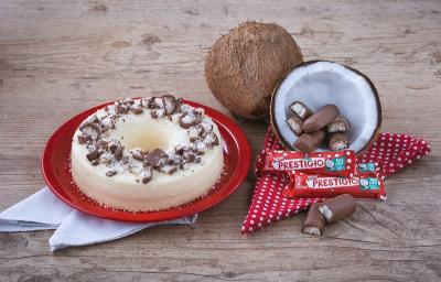 Foto em tons de vermelho e marrom da receita de pudim de prestígio servida em um prato de porcelana vermelho com panos brancos embaixo. Ao lado, um coco cortado ao meio com prestígios dentro e mais embalagens a frente, além de um pano vermelho com bolas Foto em tons de vermelho e marrom da receita de pudim de prestígio servida em um prato de porcelana vermelho com panos brancos embaixo. Ao lado, um coco cortado ao meio com prestígios dentro e mais embalagens a frente, além de um pano vermelho com bolas