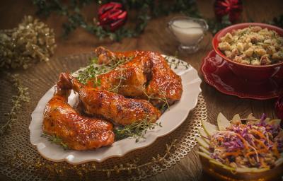 Fotografia em tons de vermelho em uma bancada de madeira escura, um prato oval branco ao centro com as sobrecoxas de frango em cima dele. Ao lado, potinhos com salada e farofa. Ao fundo, decoração e enfeites natalinos. Fotografia em tons de vermelho em uma bancada de madeira escura, um prato oval branco ao centro com as sobrecoxas de frango em cima dele. Ao lado, potinhos com salada e farofa. Ao fundo, decoração e enfeites natalinos.