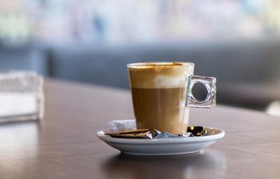 fotografia em tons de marrom e branco de uma bancada marrom vista de frente. Contém um pratinho redondo branco com uma xicara de café transparente, dentro contém café e chantilly por cima.