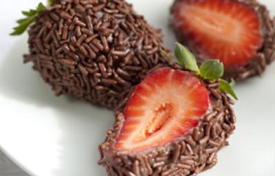 fotografia em tons de branco, marrom e vermelho tirada de um prato branco, contém 3 morangos cobertos por chocolate e chocolate granulado. Dois deles estão pela metade mostrando o morango
