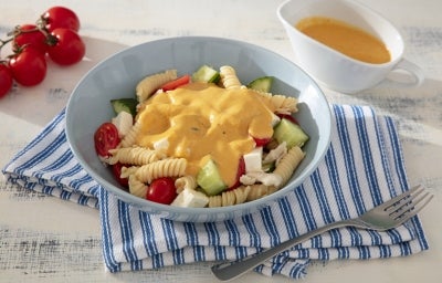 Fotografia mostra um prato azul com fusilli, cubos de vegetais, frango e tomates cereja, tudo envolvido em um molho amarelo cremoso, com cores vibrantes em destaque e textura suculenta.