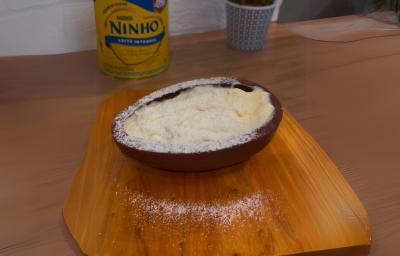 Fotografia em tons de marrom claro. No centro da imagem existe uma casca de ovo recheada com uma mousse de leite NINHO e polvilhada com leite NINHO. Ao fundo existe uma lata de leite NINHO.