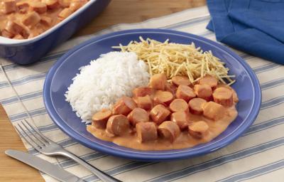 Fotografia em tons de azul em uma bancada de madeira com um jogo americano branco com listras azuis, um prato fundo azul com o strogonoff de salsicha, arroz branco e batata palha. Ao fundo, um recipiente com mais strogonoff de salsicha. Fotografia em tons de azul em uma bancada de madeira com um jogo americano branco com listras azuis, um prato fundo azul com o strogonoff de salsicha, arroz branco e batata palha. Ao fundo, um recipiente com mais strogonoff de salsicha.