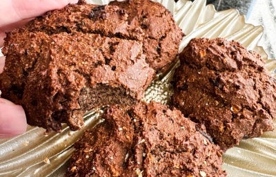 Fotografia em tons claros de uma mão feminina segurnaod um cookie fit, grande, de nescau. No fundo tem um prato raso dourado com mais unidades do cookie.