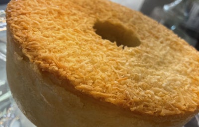 Bolo de leite com furo central e cobertura dourada de coco ralado, apoiado em suporte de vidro.