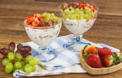 Fotografia em tons de azul e bege em uma bancada e madeira escura, um pano listrado em azul e bege, duas taças de vidro com a sobremesa de creme com morangos e uvas picados. Ao lado, um potinho com morangos em cima e cachos de uvas. Fotografia em tons de azul e bege em uma bancada e madeira escura, um pano listrado em azul e bege, duas taças de vidro com a sobremesa de creme com morangos e uvas picados. Ao lado, um potinho com morangos em cima e cachos de uvas.