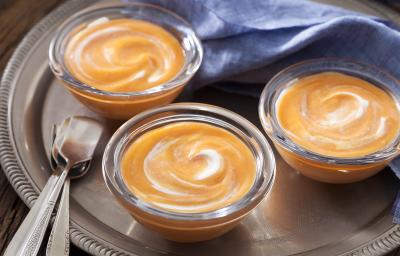 Fotografia em tons de prata e laranja de uma bandeja prata com três potinhos de vidro com o doce de abóbora cremoso. Ao fundo um paninho azul, a frente duas colheres de metal. Fotografia em tons de prata e laranja de uma bandeja prata com três potinhos de vidro com o doce de abóbora cremoso. Ao fundo um paninho azul, a frente duas colheres de metal.