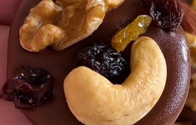 Fotografia em tons de marrom com várias moedinhas de chocolate ao centro enfeitadas com frutas secas, nozes e castanha de caju