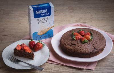 Foto da receita de bolo cremoso de aveia servida em um prato disforme branco com outro prato ao lado servido de uma fatia do mesmo bolo, tudo isso sobre um pano cor de rosa em cima de uma mesa de madeira Foto da receita de bolo cremoso de aveia servida em um prato disforme branco com outro prato ao lado servido de uma fatia do mesmo bolo, tudo isso sobre um pano cor de rosa em cima de uma mesa de madeira