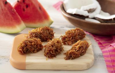 Foto da receita de cocada com casca de melancia. Observa-se uma tábua de madeira com 5 cocadas dispostas. Atrás, uma decoração com pedaços de melancia e pedaços de coco