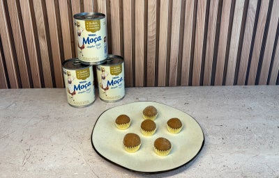 Seis brigadeiros em prato claro diante de três latas de LEITE MOÇA® sobre bancada e painel de madeira.