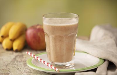 Fotografia em tons de cinza de uma bancada cinza com um prato branco com borda verde sobre ele um canudo listrado branco e vermelho e um copo de vidro com a vitamina de banana e maçã. Ao fundo um cacho de banana, uma maçã e um paninho cinza. Fotografia em tons de cinza de uma bancada cinza com um prato branco com borda verde sobre ele um canudo listrado branco e vermelho e um copo de vidro com a vitamina de banana e maçã. Ao fundo um cacho de banana, uma maçã e um paninho cinza.