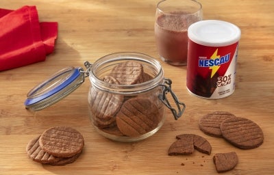 Fotografia de um potinho de vidro com tampa sobre uma mesa de madeira clara, ao lado da lata de NESCAU Extra Cacau 30% e um copo de vidro com leite e nescau. Dentro e em volta do pote tem os biscoitinhos amanteigados de nescau. Fotografia de um potinho de vidro com tampa sobre uma mesa de madeira clara, ao lado da lata de NESCAU Extra Cacau 30% e um copo de vidro com leite e nescau. Dentro e em volta do pote tem os biscoitinhos amanteigados de nescau.