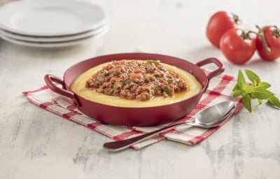 Imagem de uma caçarola vermelha com polenta e carne moída sobre um paninho xadrez vermelho e branco. Compondo a imagem, uma colher, manjericão, tomates e pratos brancos.